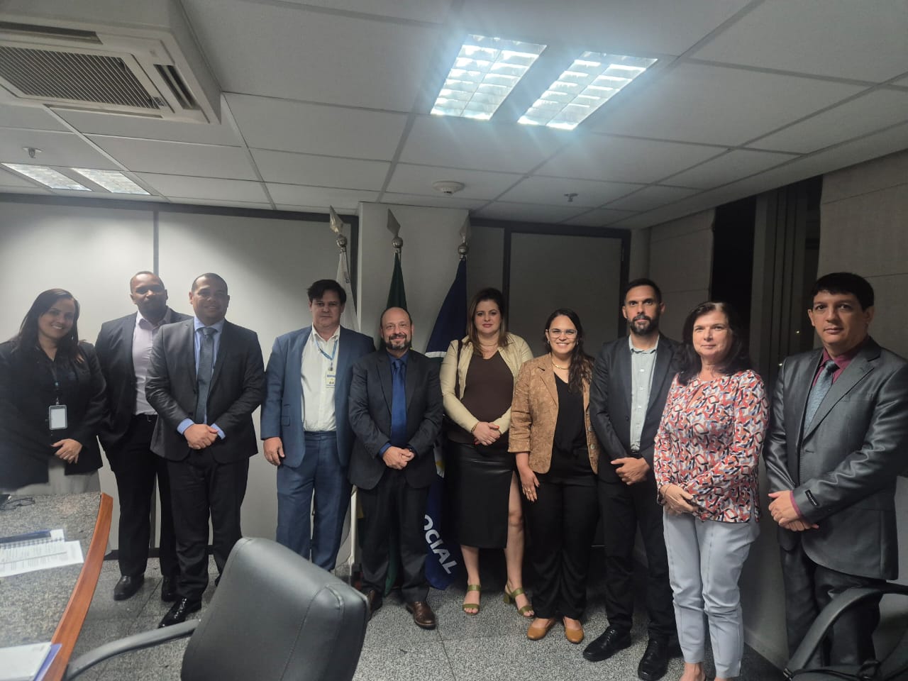Grupo de 11 pessoas em traje formal posando para foto em sala de reuniões, com bandeiras institucionais ao fundo.
